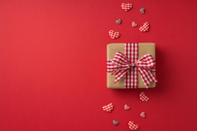 Top view photo of st valentine's day decorations kraft paper giftbox with checkered ribbon bow and hearts on isolated red background with blank space