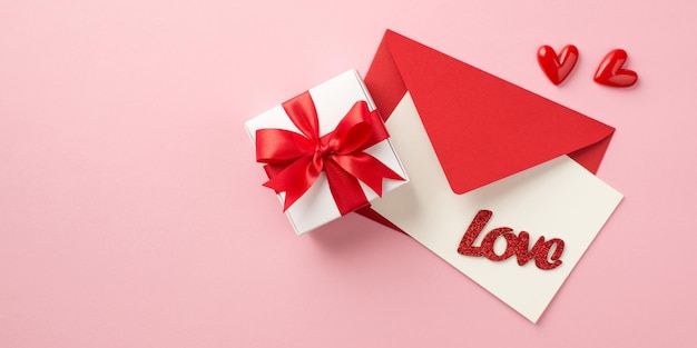 Top view photo of st valentine's day decorations red envelope with card and inscription love white giftbox with red bow and small hearts on isolated pastel pink background with copyspace