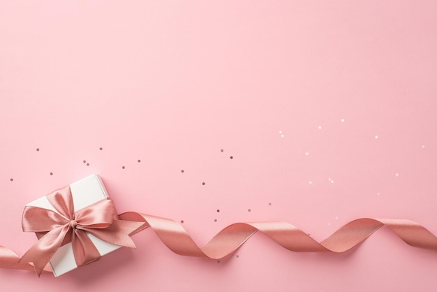 Top view photo of st valentine's day decorations white gift box with bow pink curly ribbon and sequins on isolated pastel pink background with copyspace