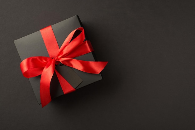 Top view photo of stylish giftbox in black packaging with vivid red satin ribbon bow and tag on isolated black background with copyspace