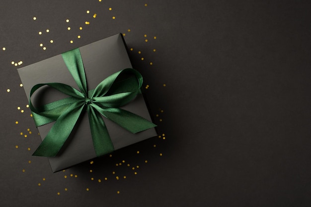 Top view photo of stylish giftbox with green ribbon bow and golden confetti on isolated black background with empty space