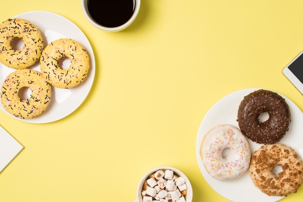 Top view photo of two cups of drink with marshmallow and coffee two plates with different color donuts and cellphone on isolated pastel yellow background with copyspace