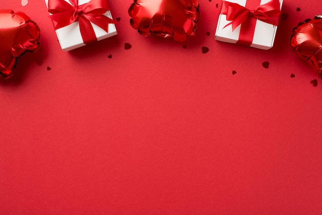 Top view photo of valentine's day decorations white gift boxes with red ribbon bows heart shaped balloons and shiny confetti on isolated red background with blank space