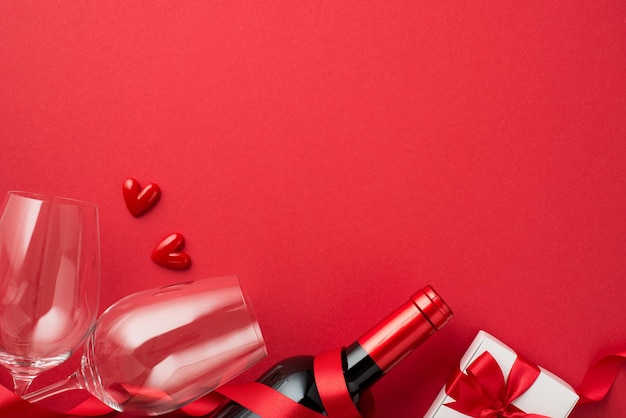 Top view photo of valentine's day decorations white giftbox with bow red ribbon small hearts two wineglasses and wine bottle on isolated red background with copyspace