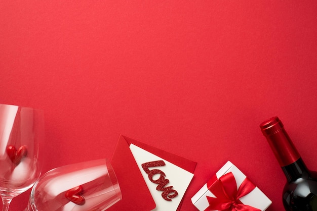 Top view photo of valentine's day decorations white giftbox with bow small hearts in two wineglasses envelope card with inscription love and wine bottle on isolated red background with empty space