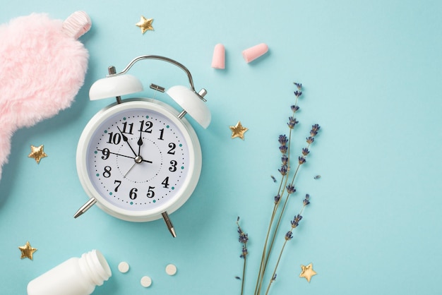 Top view photo of white alarm clock sprig of lavender pink fluffy sleeping mask earplugs open bottle with pills and golden stars on isolated pastel blue background