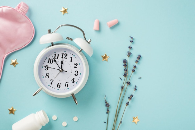 Top view photo of white alarm clock sprig of lavender pink silk sleeping mask pink earplugs open bottle with pills and golden stars on isolated pastel blue background