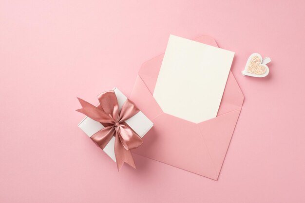 Top view photo of white giftbox with pink silk ribbon bow decorative heart and open pink envelope with card on isolated pastel pink background with copyspace