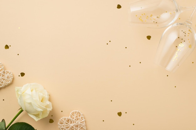 Top view photo of white rose two wineglasses with golden sequins heart shaped confetti and rattan hearts on isolated beige background with empty space