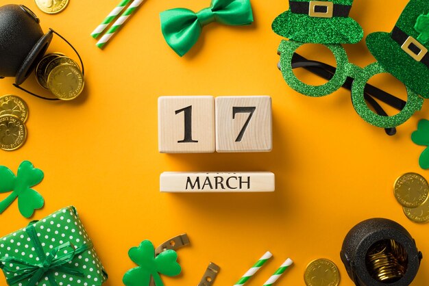 Top view photo of wooden cubes calendar 17 march st patricks day decor hat shaped party glasses straws green bowtie giftbox horseshoe shamrocks pots and gold coins on isolated yellow background