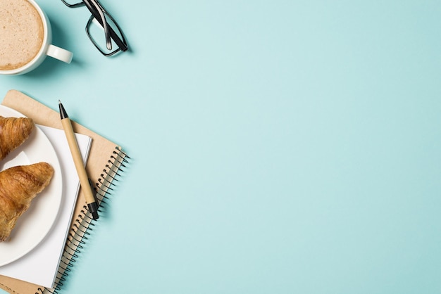 Top view photo of workplace glasses cup of frothy coffee pen planners and plate with two croissants on isolated light blue background with copyspace