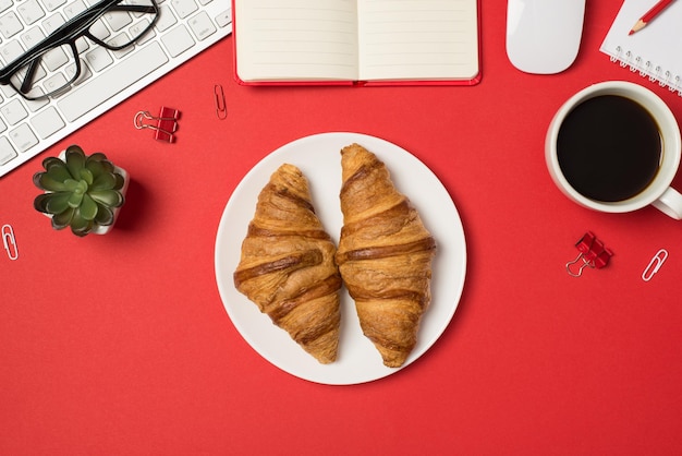 Top view photo of workplace keyboard glasses binder clips flowerpot mouse pencil planners plate with two fresh croissants and cup of coffee on isolated red background