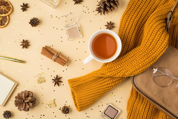 Top view photo of yellow sweater leather purse binder clips pen notebook cup of tea eyeshadows glasses pine cones anise dried lemon slices cinnamon golden sequins on isolated light beige background