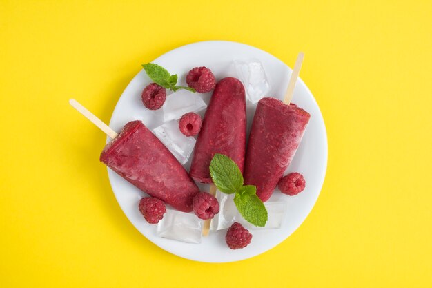 Photo top view of popsicles with  raspberry on the white plate on the yellow background. copy space.