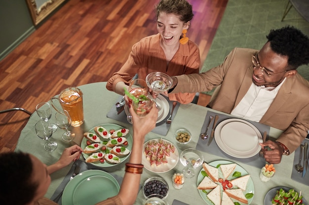 Top view portrait of elegant mixed-race couple enjoying dinner with friends indoors and clinking glasses over table, copy space