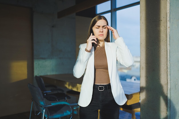 Top view of positive woman in casual clothes standing near big window and talking on phone giving information during remote project Modern workspace for remote work