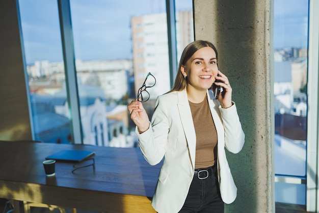 Top view of positive woman in casual clothes standing near big window and talking on phone giving information during remote project Modern workspace for remote work