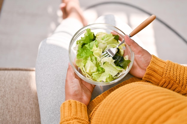 Top view of a pregnant woman holding a salad bowl eating salad on sofa in her living room