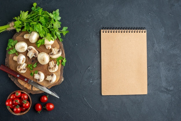 Top view of raw mushrooms and greens knife on wooden board white towel and fresh tomatoes in a bowl spiral notebook on black background with free space