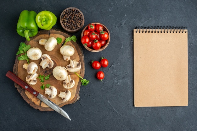 Top view of raw mushrooms and greens knife on wooden board white towel and fresh vegetables spiral notebook on black background