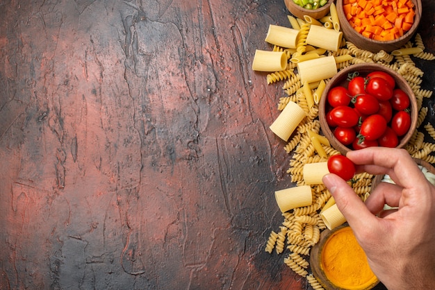 top view raw pastas penne rigatoni spirals chopped vegetables and turmeric in bowls cherry tomato in female hand on wooden table