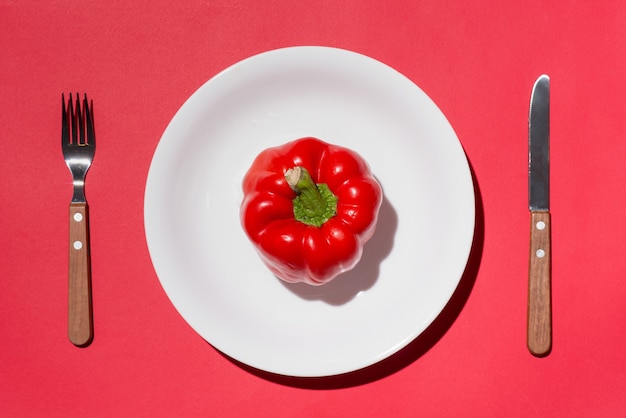 Top view of red bell pepper on white plate with knife and fork on red background