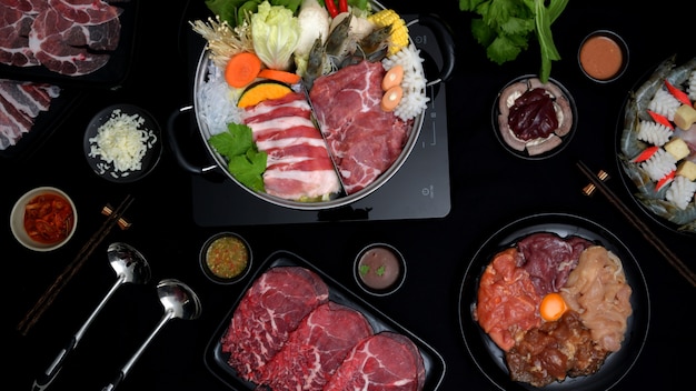 Top view of Shabu-Shabu in hot pot, fresh sliced meat, sea food, vegetables and dipping sauce with black background