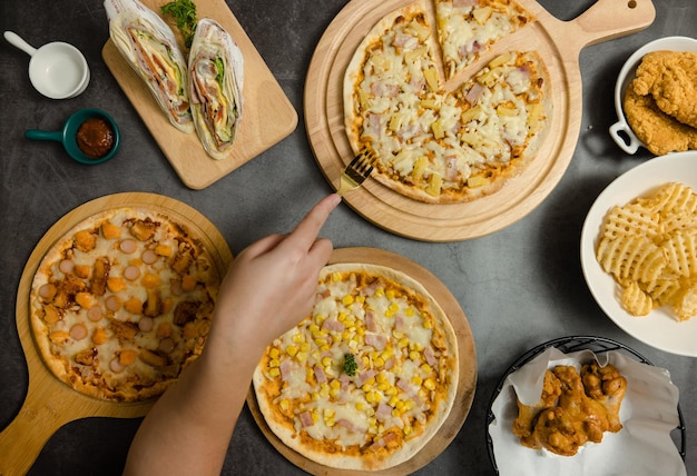 Top view shot of various kinds of delicious tasty juicy cheesy pizza placed on wooden board on party table around with other fast food while unrecognizable people picking up eating dinner together