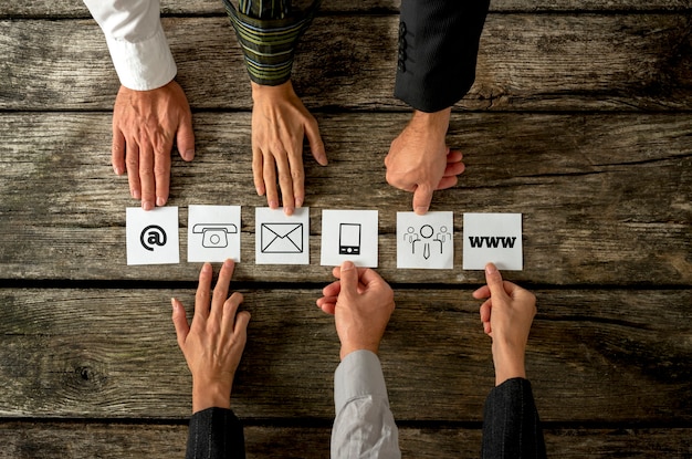 Top view of six people placing white cards with various contact icons in a row on a textured wooden desk.