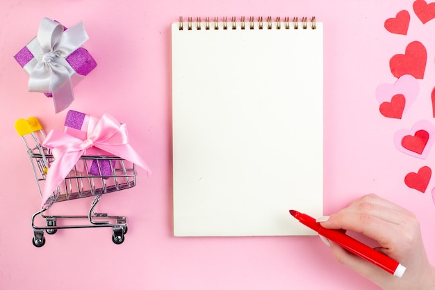 top view small gifts red marker in female hand mini market trolley with gift notebook on pink background