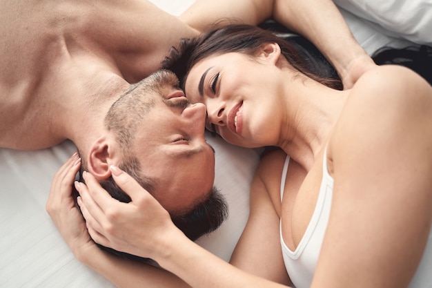 Top view of a smiling sleepy serene dark-haired woman lying in bed with her husband