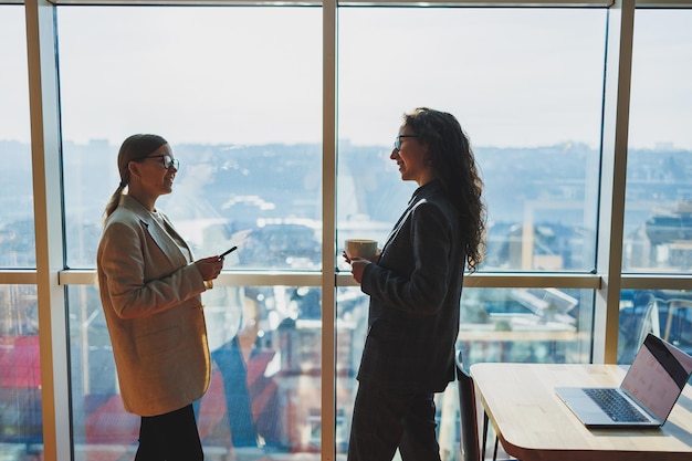 Top view of smiling young confident business workers in casual clothes standing on modern office background and talking during day break