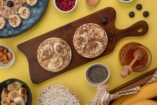 Top view of snacks with banana peanut butter sesame on crispbread on cutting board with red currant almond jam blackthorn on yellow background