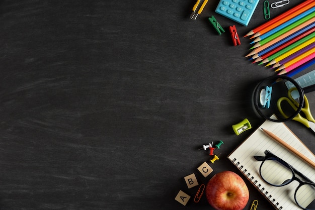 Top view of stationery or school supplies with books, color pencils, calculator, laptop, clips and red apple on chalkboard background.