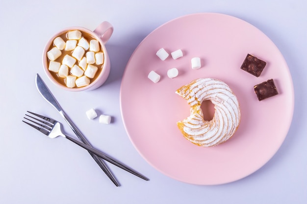 Top view still life of a bitten cake on a pink plate, cutlery and a cup of cocoa with marshmallows. Selective focus, horizontal orientation.