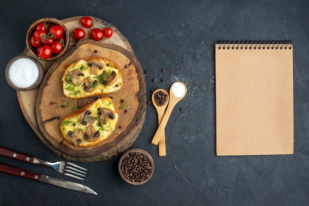 Top view of tasty snack with mushrooms tomatoes salt on wooden board cutlery set notebook on black background