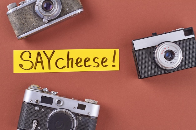 Top view three retro photo cameras on red background say cheese handwriting on yellow paper