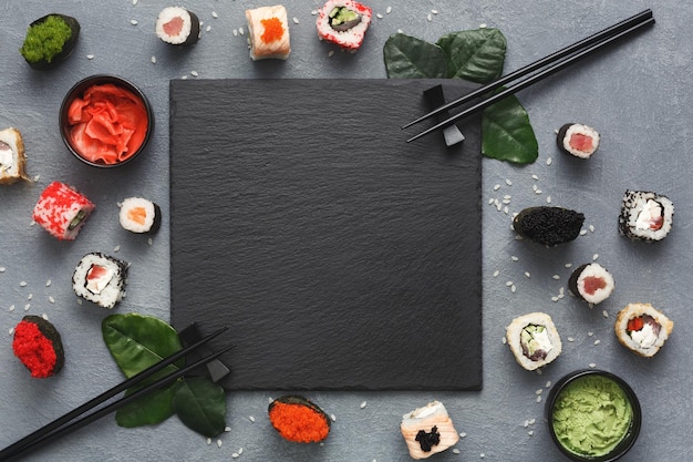 Top view on traditional japanese empty square black slate framed with sushi, ginger, soy sauce, wasabi bowls, green leaves and chopsticks on rustic grey background, copy space