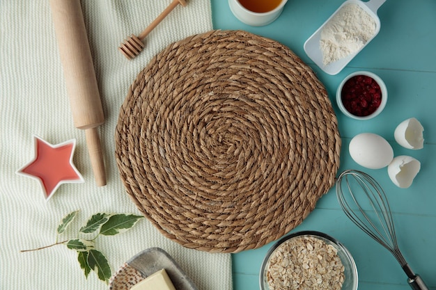 Top view of trivet for hot dishes with jam butter oat red currant egg flour egg shell rolling pin whisk on cloth on blue background