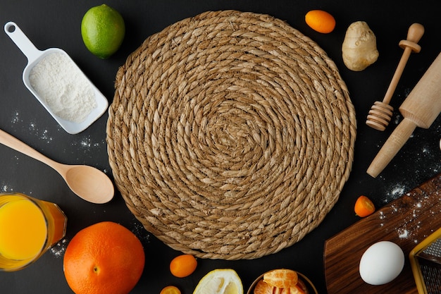 Top view of trivet with egg lemon lime orange tangerine kumquat flour ginger orange juice with spoon honey dipper and rolling pin on black background