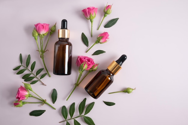 Top view of two glass bottles with cosmetics face serum against the background of pink rose buds and green leaves