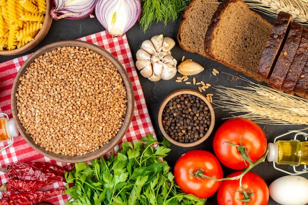 Top view of uncooked buckwheat in a brown bowl on towel fresh vegetables fallen oil bottle salt green bundle bread slices