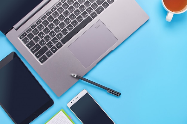 Top view upon gadgets on blue background, the composition of a laptop, white headphones, phone, glass with a drink and car keys