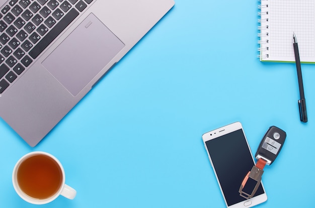 Top view upon gadgets on blue background, the composition of a laptop, white headphones, phone, glass with a drink and car keys