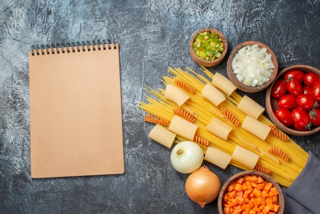 top view various pastas rigatoni spaghetti spirals notebook chopped vegetables in bowls on grey background