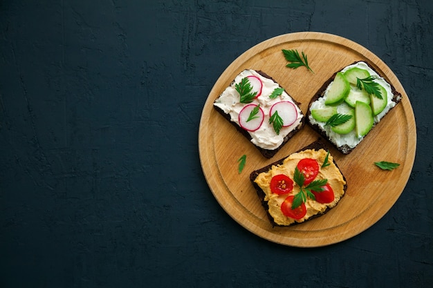 Top view vegetarian rye bread toasts with cottage cheese, hummus, avocado, raddish and tomato on a wooden board, black background, copy-space