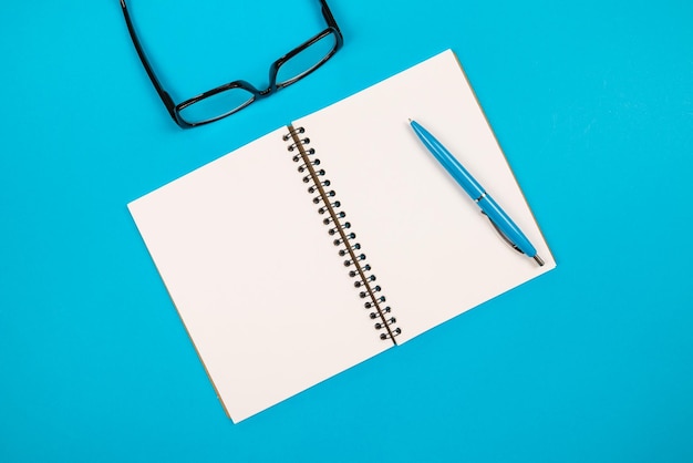 Top view of white blank notepad with pencil and glasses on isolated blue background with copy space