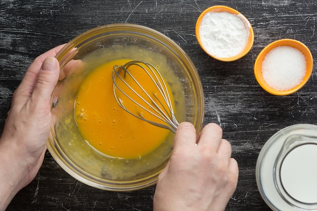 Top view of woman hands mixing with whisk yolks in bowl for making custard cream on the black background