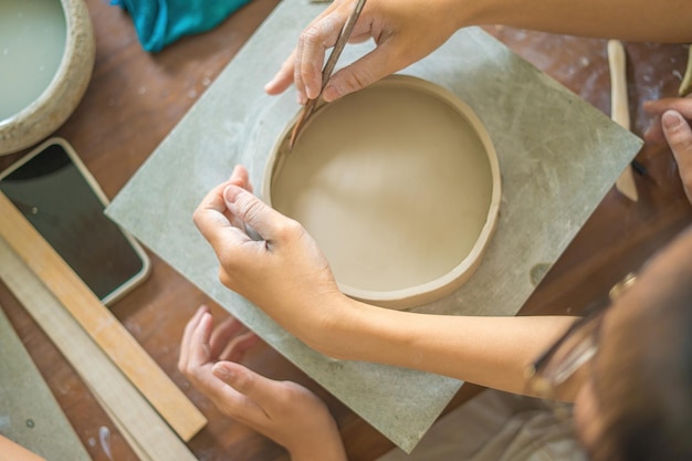 Top view Woman potter working on potters wheel making ceramic pot from clay in pottery workshop Focus hand young woman attaching clay product part to future ceramic product Pottery workshop