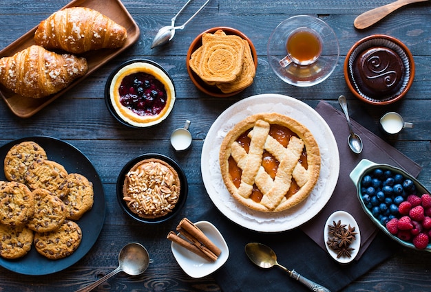 Top view of a wood table full of cakes, fruits, coffee, biscuits, spices and more